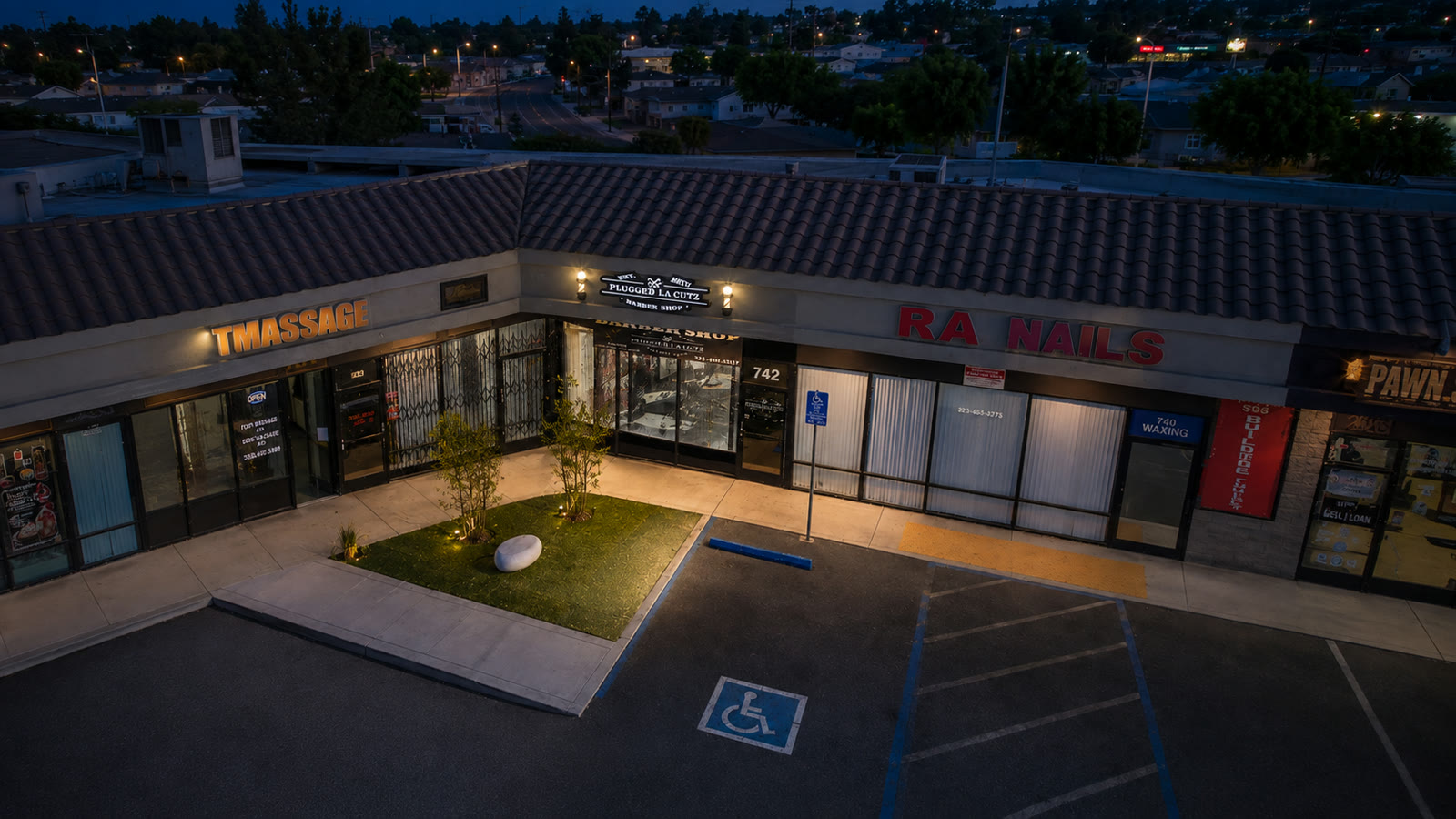 Evening drone view of the plaza — the lit Plugged LA Cutz sign marks suite 742.