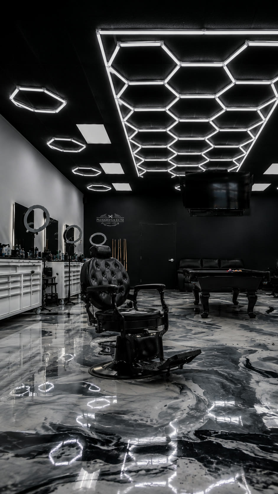 An empty black tufted barber chair on the marble floor, waiting under the hex lights.